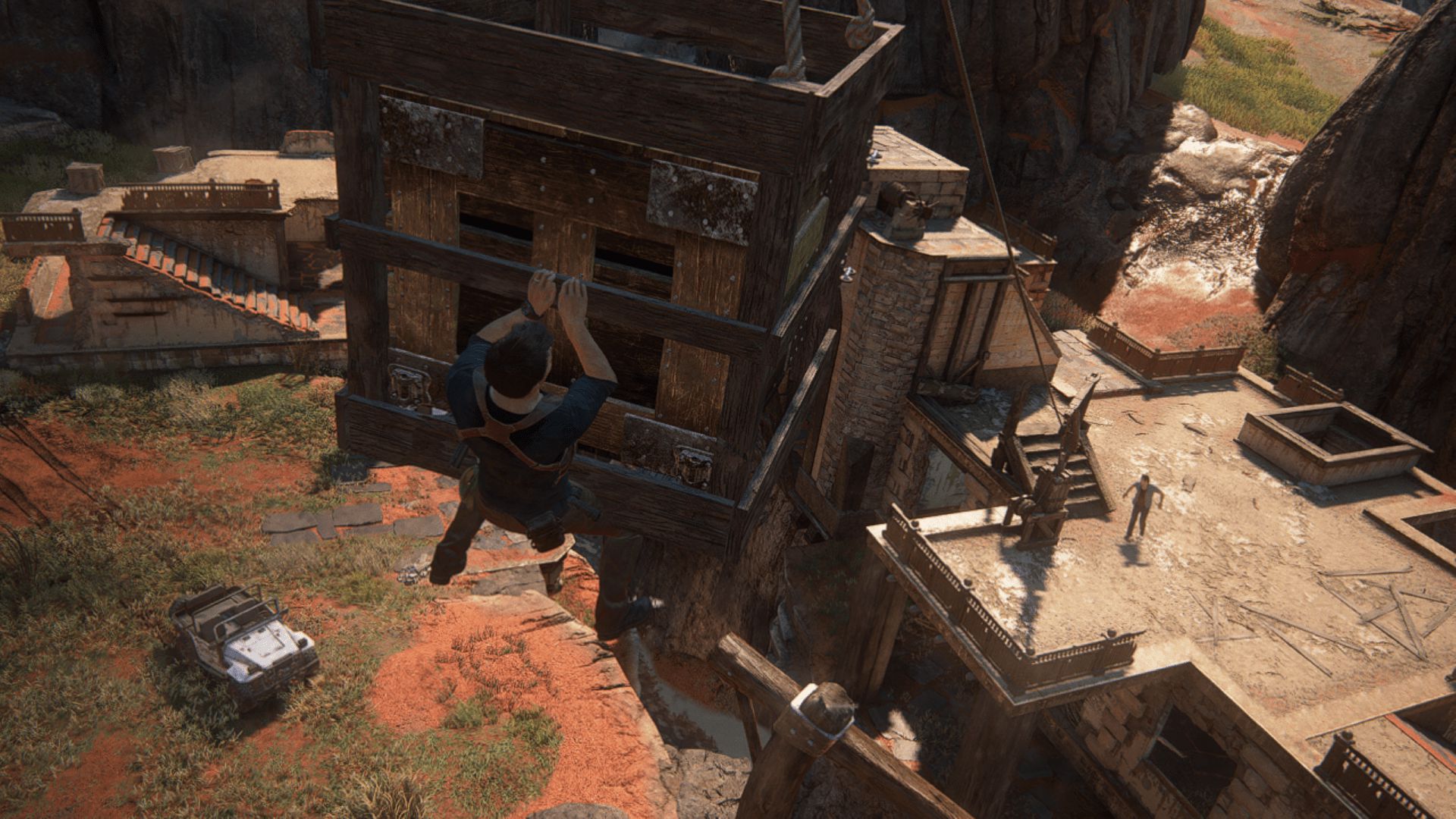 A man clings to a suspended wooden crate above a ruin, while another figure stands on a rooftop below.