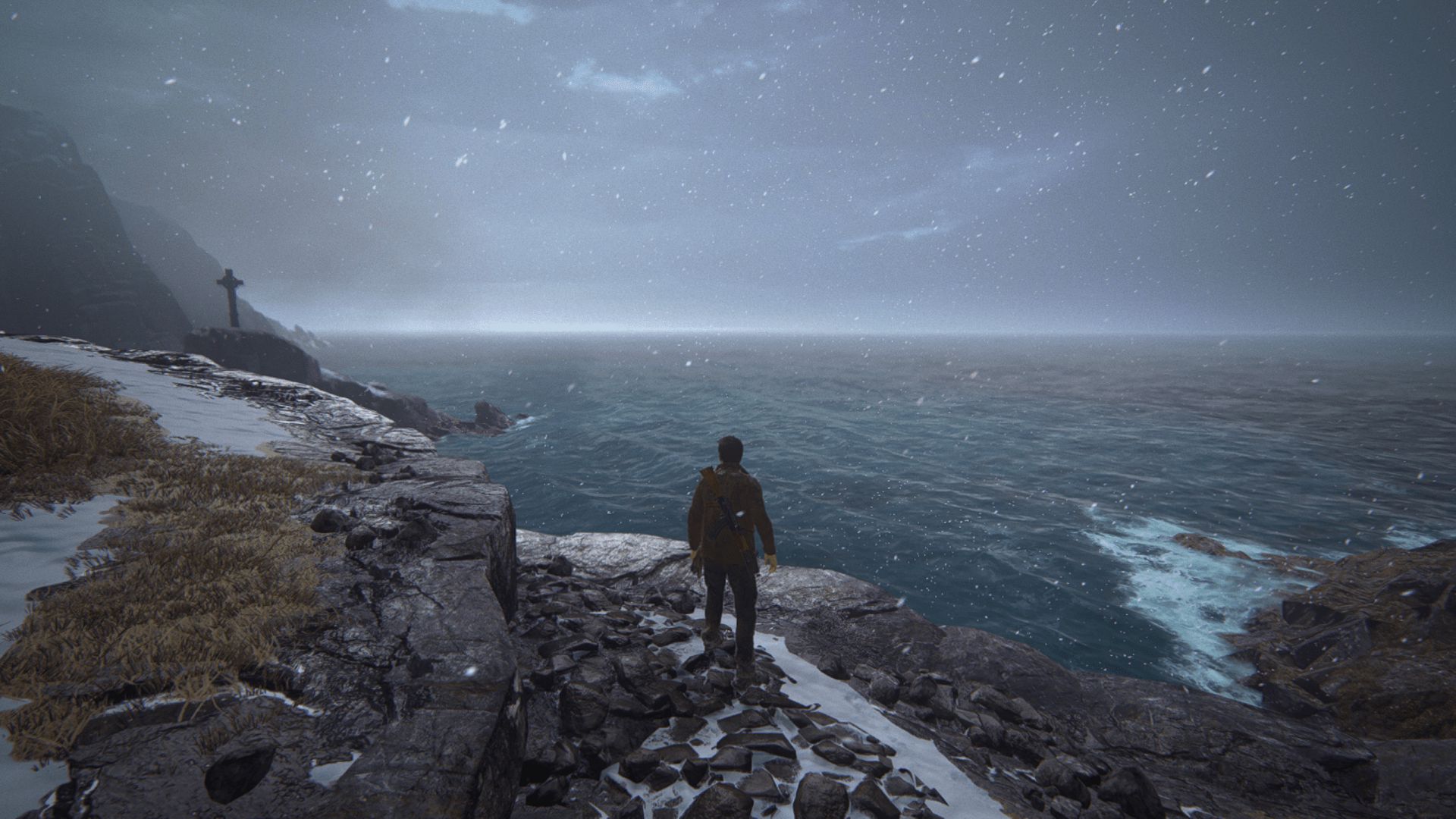 A man stands on a rocky, snow-dusted cliff edge overlooking a stormy ocean, with snow falling and a large stone cross visible in the distance.